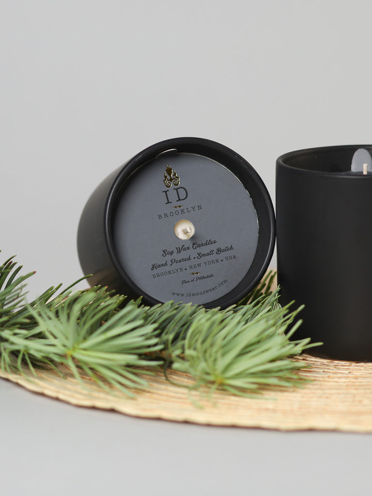 A black SSC - Sultry Santal and Cedarwood Candle by ID Menswear, with a burning wick, emits a cedarwood scent next to a pine-decorated round mat. Another holder is on its side, showing the ID Menswear label and crafted from luxurious soy wax.